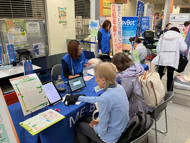 屋内イベント会場で、スタッフが来場者にME-BYOに関する測定案内をしている様子