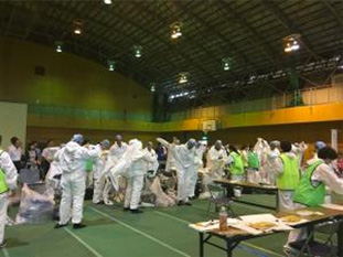 体育館のような広い空間で活動する白い防護服を着た人物たち